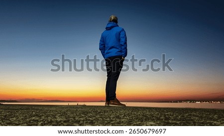 From low angle, man in a blue jacket stands alone on pier, looking out at distant city lights across the water during the tranquil blue hour after sunset, evoking a sense of calm and urban solitude.