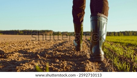 Low angle: man walking in rubber boots in a farmer's field, the blue sky above the horizon. Man walking through an agricultural field. Farmer walks through a plowed field in early spring. 