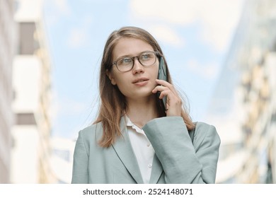 Low angle medium closeup of young Caucasian female manager wearing eyeglasses having phone call while walking outdoors