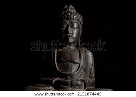 Low angle, medium shot, horizontal image of a sitting buddha statue in lotus position ina a black background. It is made of palm wood with dull, dark brown colors. Dark image, copy space.