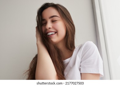 Low angle of merry female teenager in white t shirt touching long hair and smiling with closed eyes, near window in light room at home - Shutterstock ID 2177027503