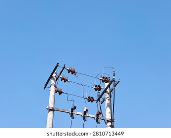 A low angle minimalist shot of double concrete utility poles with electrical ceramic insulators and cables against a clear blue sky. A detailed view of double concrete electricity poles equipped with rows of brown porcelain insulators and fuses under a bright daytime sky.