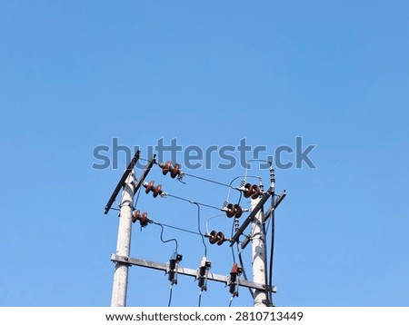 A low angle minimalist shot of double concrete utility poles with electrical ceramic insulators and cables against a clear blue sky. A detailed view of double concrete electricity poles equipped with rows of brown porcelain insulators and fuses under a bright daytime sky.