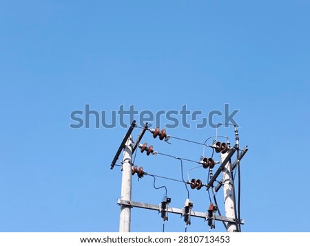 A low angle minimalist shot of double concrete utility poles with electrical ceramic insulators and cables against a clear blue sky. A detailed view of double concrete electricity poles equipped with rows of brown porcelain insulators and fuses under a bright daytime sky.