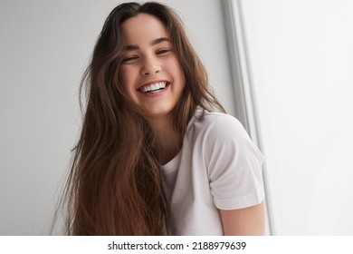 Low angle of optimistic young female millennial with long brown hair in t shirt, smiling brightly and looking at camera against white wall at home - Shutterstock ID 2188979639