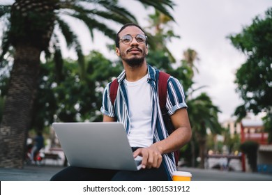 Low angle of pensive male remote worker in casual summer outfit and eyeglasses browsing laptop and looking away on city street