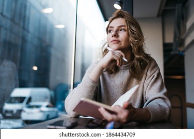 Low angle of pensive young female writer looking away and thinking while working on new article sitting in modern cafe with notebook and smartphone - Shutterstock ID 1864434709