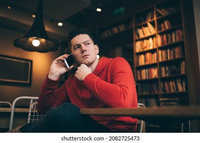 Low Angle Of Pensive Young Guy In Red Hoodie Sitting With Hand At Chin While Making Phone Call And Looking Away