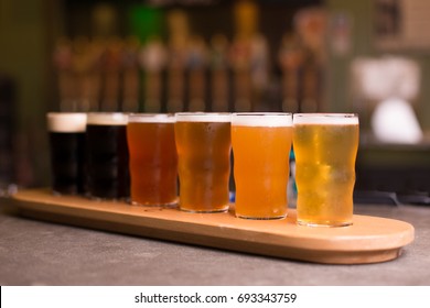 Low Angle Perspective Close Up Of Craft Beer Tasting Flight At Local Brewery Of Small Pint Glasses In Row On Wooden Tray With Rainbow Variety Of Golden Yellow Hoppy Ales To Dark Malt Stouts On Bar