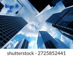 A low angle perspective looking directly upward at a cluster of high rise skyscrapers in a dense urban business district creates a striking geometric composition against the sky The buildings are predominantly constructed of glass and steel featuring a variety of architectural styles and surface textures ranging from smooth reflective glass panels to complex grid patterns formed by window frames and structural supports On the left a light colored building displays a prominent and repetitive rectangular grid of windows that recedes into the distance while the center and right foreground are occupied by skyscrapers with dark blue reflective glass facades that mirror the surrounding structures and the sky above The sky visible in the central opening between the towering structures is a vibrant blue filled with soft white and grey clouds that are brightly illuminated by natural daylight A single street lamp on a tall thin pole extends into the frame from the upper left adding a functional urban element to the architectural scene The cool blue color palette of the image is enhanced by the reflections on the glass surfaces which create a sense of depth and atmospheric perspective as the buildings seem to converge toward a single point in the sky Every detail of the building facades including the individual window panes and the sharp edges of the corners is clearly defined in the bright outdoor light The composition highlights the massive scale and verticality of modern city life emphasizing the intersection of engineering and design in a metropolitan setting where light and shadow play across the varying materials of the urban landscape. 