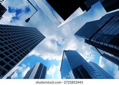 A low angle perspective looking directly upward at a cluster of high rise skyscrapers in a dense urban business district creates a striking geometric composition against the sky The buildings are predominantly constructed of glass and steel featuring a variety of architectural styles and surface textures ranging from smooth reflective glass panels to complex grid patterns formed by window frames and structural supports On the left a light colored building displays a prominent and repetitive rectangular grid of windows that recedes into the distance while the center and right foreground are occupied by skyscrapers with dark blue reflective glass facades that mirror the surrounding structures and the sky above The sky visible in the central opening between the towering structures is a vibrant blue filled with soft white and grey clouds that are brightly illuminated by natural daylight A single street lamp on a tall thin pole extends into the frame from the upper left adding a functional urban element to the architectural scene The cool blue color palette of the image is enhanced by the reflections on the glass surfaces which create a sense of depth and atmospheric perspective as the buildings seem to converge toward a single point in the sky Every detail of the building facades including the individual window panes and the sharp edges of the corners is clearly defined in the bright outdoor light The composition highlights the massive scale and verticality of modern city life emphasizing the intersection of engineering and design in a metropolitan setting where light and shadow play across the varying materials of the urban landscape. 