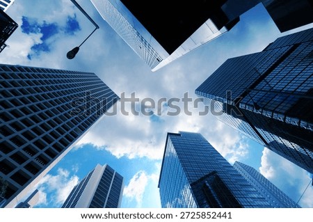 A low angle perspective looking directly upward at a cluster of high rise skyscrapers in a dense urban business district creates a striking geometric composition against the sky The buildings are predominantly constructed of glass and steel featuring a variety of architectural styles and surface textures ranging from smooth reflective glass panels to complex grid patterns formed by window frames and structural supports On the left a light colored building displays a prominent and repetitive rectangular grid of windows that recedes into the distance while the center and right foreground are occupied by skyscrapers with dark blue reflective glass facades that mirror the surrounding structures and the sky above The sky visible in the central opening between the towering structures is a vibrant blue filled with soft white and grey clouds that are brightly illuminated by natural daylight A single street lamp on a tall thin pole extends into the frame from the upper left adding a functional urban element to the architectural scene The cool blue color palette of the image is enhanced by the reflections on the glass surfaces which create a sense of depth and atmospheric perspective as the buildings seem to converge toward a single point in the sky Every detail of the building facades including the individual window panes and the sharp edges of the corners is clearly defined in the bright outdoor light The composition highlights the massive scale and verticality of modern city life emphasizing the intersection of engineering and design in a metropolitan setting where light and shadow play across the varying materials of the urban landscape. 