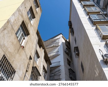 A low angle perspective looking up from a narrow alleyway at densely packed, aging multi-story residential buildings with barred windows against a clear blue sky. The scene represents dense urban living, aging city infrastructure, overpopulation, and neighborhood communities.