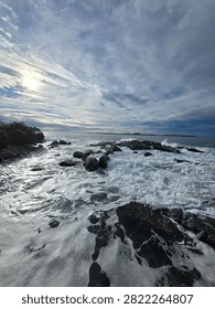 Low angle perspective looking over dark coastal rocks washed by white ocean waves under cloudy sky.