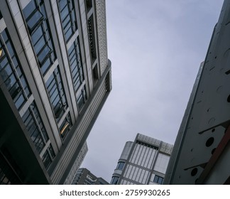 Low angle perspective looking straight up at tall modern glass and concrete office buildings converging against an overcast grey sky. The scene represents corporate environments, dense urban architecture, upward growth, and metropolitan business districts.