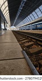 Low angle perspective of a modern train station platform with railway tracks under a curved glass canopy roof and a blue passenger train in the background. View from the edge of a concrete platform showing heavy railway sleepers and tracks inside a futuristic arched terminal with blue train cars parked alongside. Empty railway station platform under a massive arched blue roof with a passenger train waiting on the tracks during a bright day in the city.