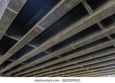 Low angle perspective view of heavy concrete bridge girders, beams, and structural support framework underneath an elevated highway overpass infrastructure.