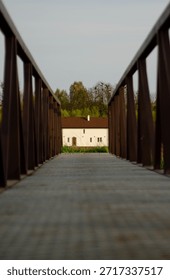 Low angle perspective view of a metal footbridge with railings leading to a white rural house. Symmetrical walkway composition symbolizing the path home, real estate access, or journey to a destinatio