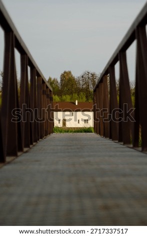 Low angle perspective view of a metal footbridge with railings leading to a white rural house. Symmetrical walkway composition symbolizing the path home, real estate access, or journey to a destinatio