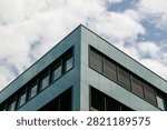 Low angle perspective view of the sharp corner of a contemporary office building featuring dark green metallic panel facade and glass windows against a blue sky with white clouds in Rotkreuz, Switzerland. Concept of modern corporate real estate and business hub