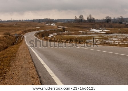 Low angle photo of tarmac curvy road at vintage brown meadows at early spring. Melancholic winding drive at countryside from left. Twisty gray asphalt path near Ergli, Latvia during rainy day. 