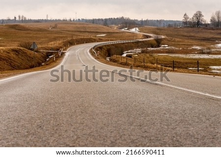 Low angle photo of tarmac road curving through vintage brown meadows at early spring. Life is not a straight road – melancholic wavy drive at countryside. Curvy gray asphalt path near Ergli, Latvia