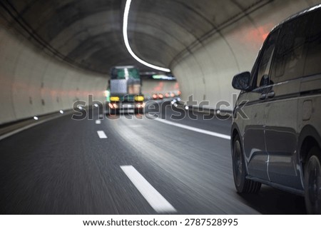 A low angle point of view shot from a moving vehicle inside a modern underground concrete tunnel, showing a gray passenger van and a blurry truck ahead with motion blur.