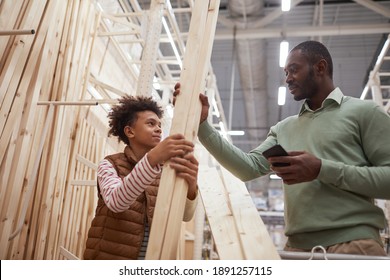 Low Angle Portrait Of African-American Father And Son Shopping Together In Hardware Store, Focus On Man Choosing Wooden Boards For Construction Or Home Improvement