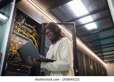 Low angle portrait of African-American female network engineer standing by server cabinet and holding laptop while working with supercomputer in data center, copy space - Powered by Shutterstock