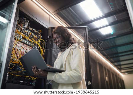 Low angle portrait of African-American female network engineer standing by server cabinet and holding laptop while working with supercomputer in data center, copy space