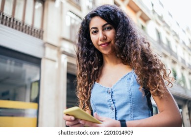 Low Angle Portrait Of An Attractive Dark Haired Woman Looking At Camera While Holding Her Phone