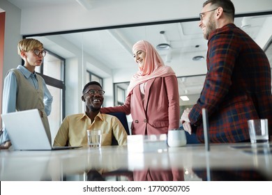 Low Angle Portrait Of Cheerful Multi-ethnic Business Team Standing By Meeting Table In Conference Room, Focus On Muslim Woman Wearing Headscarf Standing By African-American Colleague