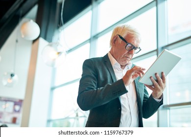 Low angle portrait of contemporary senior man using digital tablet and browsing internet while standing against window, copy space - Powered by Shutterstock