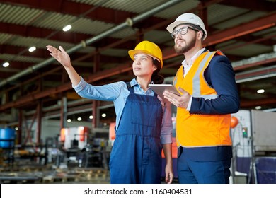 Low angle portrait of female factory worker giving tour of production workshop to businessman wearing hardhat and pointing away, copy space