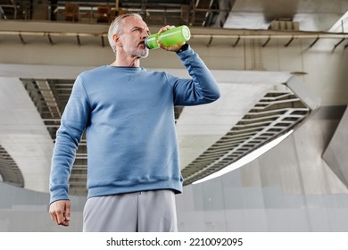 Low Angle Portrait Of Handsome Senior Man Drinking Water During Outdoor Workout In Urban Setting Under Bridge Structure, Copy Space