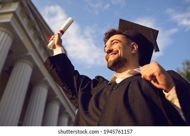 Low angle portrait of happy triumphant male graduate standing near university holding up diploma. From below of young handsome man proud of academic achievements celebrating college graduation