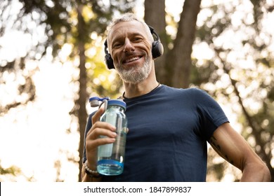 Low Angle Portrait Of Joyful Grizzled Athletic Male Drinking Water While Doing Workout In Sunny Forest