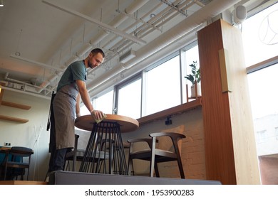 Low Angle Portrait Of Male Waiter Cleaning Tables In Cafe Or Coffee Shop While Preparing For Opening In Morning, Copy Space