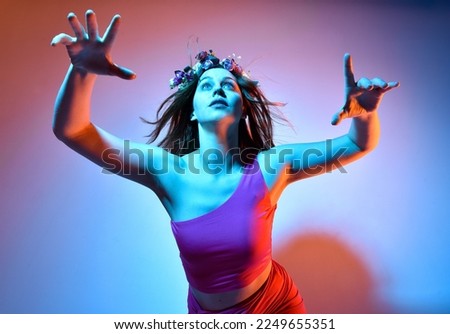 Low angle portrait of pretty girl wearing pink outfit, gestural arm poses reaching out as if casting a spell, colourful neon gel lighting, isolated on studio background.