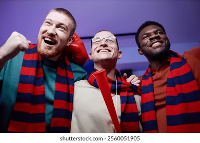 Low angle portrait of three excited sports fans huddling and cheering at home watching game match on TV and wearing matching scarves in team colors - Powered by Shutterstock