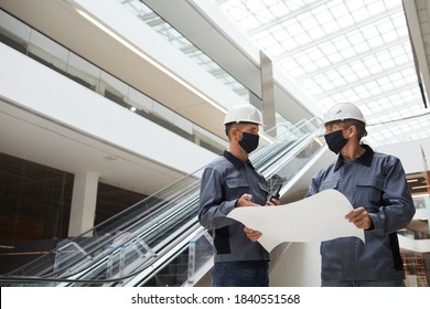 Low Angle Portrait Of Two Construction Workers Wearing Masks And Discussing Plans While Standing In Shopping Mall Or Office Building, Copy Space