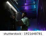 Low angle portrait of young African American data engineer working with supercomputer in server room lit by blue light and holding laptop, copy space