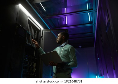 Low angle portrait of young African American data engineer working with supercomputer in server room lit by blue light and holding laptop, copy space - Powered by Shutterstock