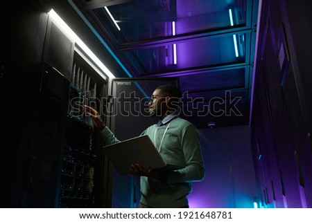 Low angle portrait of young African American data engineer working with supercomputer in server room lit by blue light and holding laptop, copy space