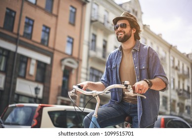 Low Angle Portrait Of Young Guy In Good Mood Riding Bike At High Speed. Copy Space In Left Side. Buildings On Background