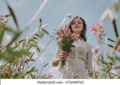 Low Angle Portrait Of Young Woman With Flower Bouquet Framed By Growing Flowers In A Field