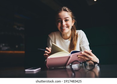 Low Angle Of Positive Female In Casual Clothes With Toothy Smile Looking At Camera Reading Interesting Book While Having Free Time