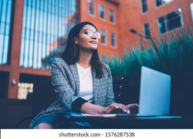 Low Angle Of Positive Freelancer Looking Away And Thinking While Typing On Laptop Keyboard While Sitting On Bench Outside Building On City Street