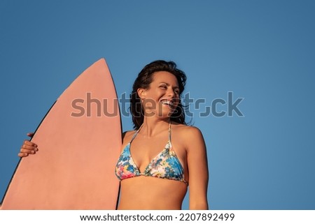 Low angle of positive Hispanic woman in bikini with surfboard standing on background of cloudless sky at sundown in summer and looking away