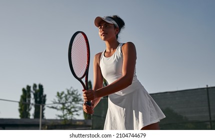 Low angle of professional African American female athlete with racket playing tennis during training on sports ground in summer evening